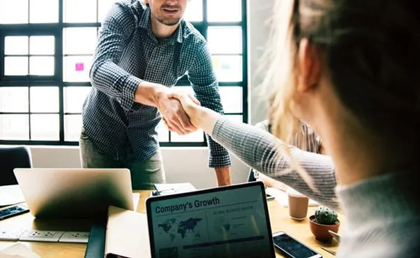 Business meeting handshake between two professionals, with laptops displaying growth metrics and tax-related documents in a modern workspace.