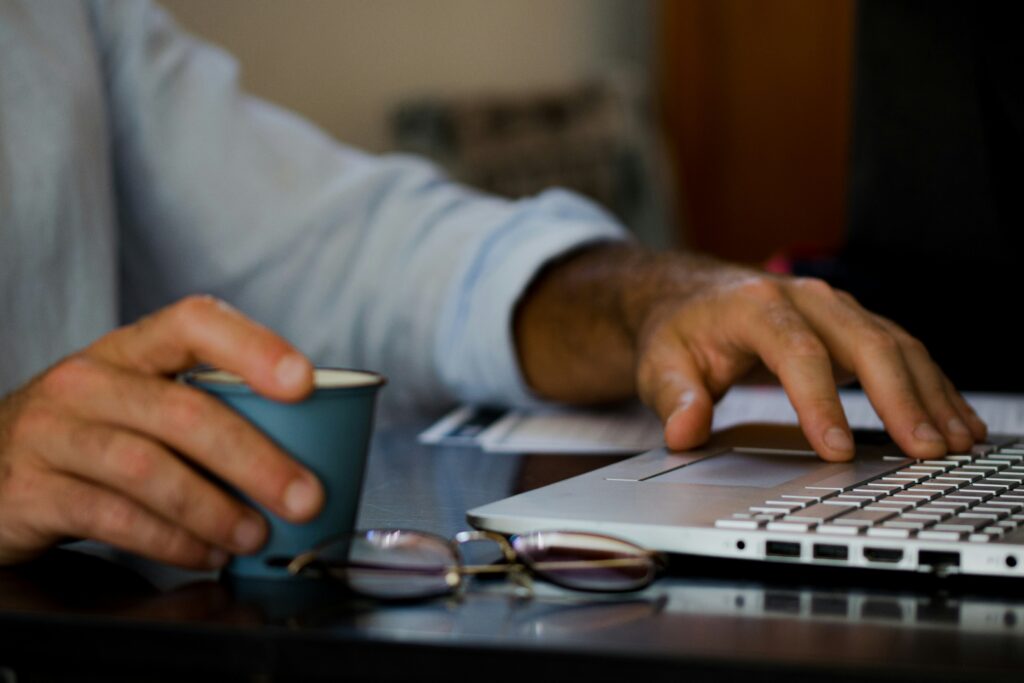 Person using a laptop to e-file 1099 NEC forms, holding a coffee cup, with reading glasses on a table.