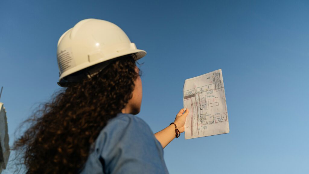 Construction professional in hard hat holding blueprints against clear sky, representing project planning and compliance in construction industry.